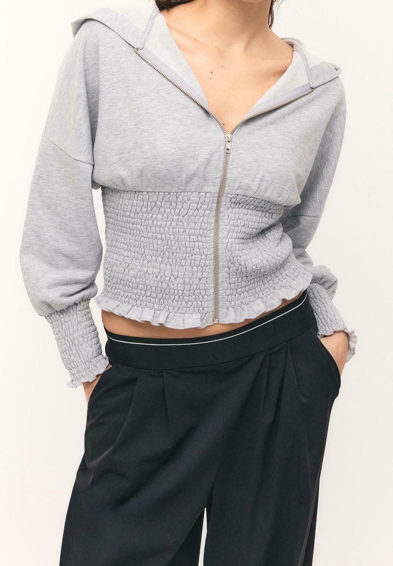 Femme portant un sweat à capuche court gris clair zippé avec taille et manches smockées, associé à un pantalon noir plissé taille haute.