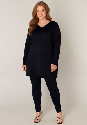 Lachende vrouw met lang haar die een zwarte tuniek met lange mouwen draagt, bijpassende legging en zwarte sandalen met bandjes tegen een effen achtergrond.