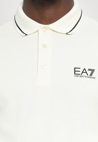 Polo-shirt in romig wit met een gestructureerde stof, drie knopen, kraag met zwarte rand en het EA7 Emporio Armani-logo op de linkerborst.