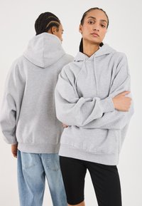 Felpa grigia realizzata in tessuto morbido, con un ampio cappuccio e spalle scese. Lo stile del modello include jeans dal taglio rilassato e pantaloni corti aderenti.