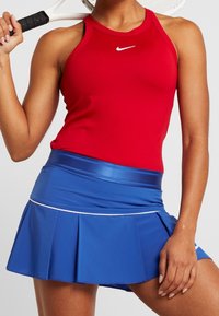 Un haut de sport sans manches rouge avec un logo blanc, associé à une jupe plissée bleue avec une ceinture contrastante et des accents blancs.