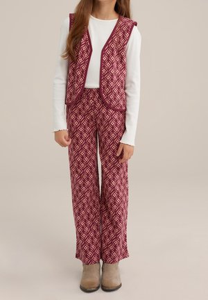 Dziewczyna ubrana w biały top z długim rękawem, pasującą bordową kamizelkę z wzorem oraz szerokie spodnie, zestawione z beżowymi botkami na neutralnym tle.