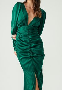 Grön satindress med långa ärmar, djup V-ringning, rynkad bodice och slits vid låret. Slät textur och åtsittande siluett.