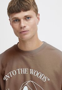 Hnědá mikina se žebrovaným výstřihem, s bílým potiskem textu "INTO THE WOODS" a ilustrovanou grafikou níže. Měkká struktura tkaniny.