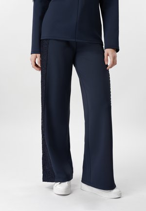 Person, die marineblaue, weite Hosen mit Spitzeneinsätzen an den Seiten und weiße Plateausneaker trägt, steht vor einem schlichten Hintergrund.