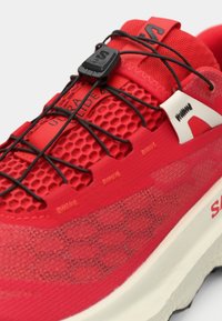 Κόκκινο αθλητικό παπούτσι με υφασμάτινο επάνω μέρος με υφή, μαύρα κορδόνια και έντονο λευκό λογότυπο. Η σόλα είναι ελαφρώς ανυψωμένη με λαστιχένια επένδυση.