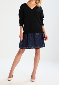 Schwarzer, flauschiger Pullover mit V-Ausschnitt und lockeren Ärmeln, kombiniert mit einem dunkelblauen, semi-transparenten Rock mit Blumenmustern. Beigefarbene Absätze.