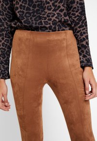 Femme portant un pantalon en daim marron et un haut à manches longues imprimé léopard noir, montrant le ventre et les mains.