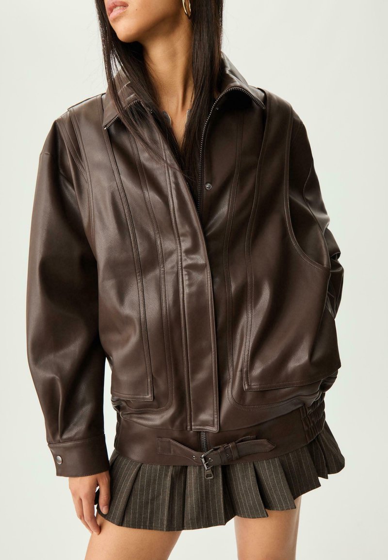 Donna che indossa un'oversized giacca in pelle marrone scuro con dettagli di cucitura e una mini gonna plissettata con cintura, su uno sfondo neutro.