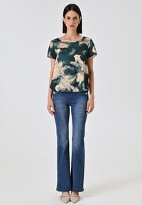Top floreale a maniche corte in teal e beige, abbinato a jeans blu a zampa d'elefante. Tessuto leggero, scollatura rotonda e dettagli di cuciture frontali visibili.