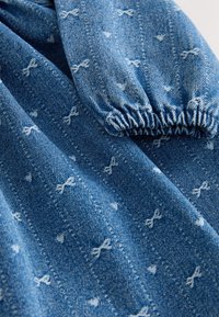 Džínovina s modrým odtieňom zdobená bielymi vzormi mašlí. Obsahuje nariasené manžety s elastickým detailom, ktoré majú jemnú textúru a dizajn.