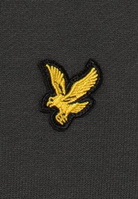 Logo brodat cu o vultur în galben și negru pe un material texturat de culoare gri închis. Vulturul are aripi desfășurate cu detalii fine.
