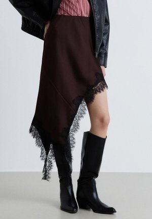 Donna con gonna asimmetrica marrone scuro con bordatura in pizzo nero, stivali neri a metà polpaccio, camicia a righe rossa e giacca di pelle nera.