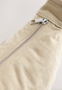 Bēšas krāsas korduro audums ar ribotu faktūru, sudraba krāsas metāla rāvējslēdzējs ar vilkšanas cilpu, novietots diagonāli. Redzama dekoratīvā šūšana.