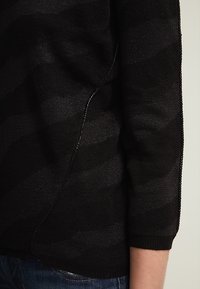 Svart stickad tröja med subtilt vågmönster, texturerad yta och kontrasterande sömmar längs sömmen. Fitted mudd och sidodetaljer synliga.