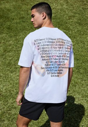 Tânăr bărbat stând pe iarbă, purtând un tricou alb cu numele orașelor și date tipărite pe spate, asortat cu pantaloni scurți negri.