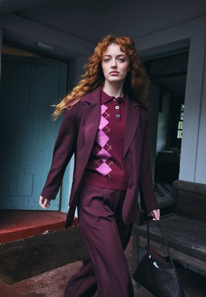 Jeune femme aux longs cheveux bouclés roux marche à l’intérieur portant un costume bordeaux et un pull à losanges rose, tenant un sac à main noir.