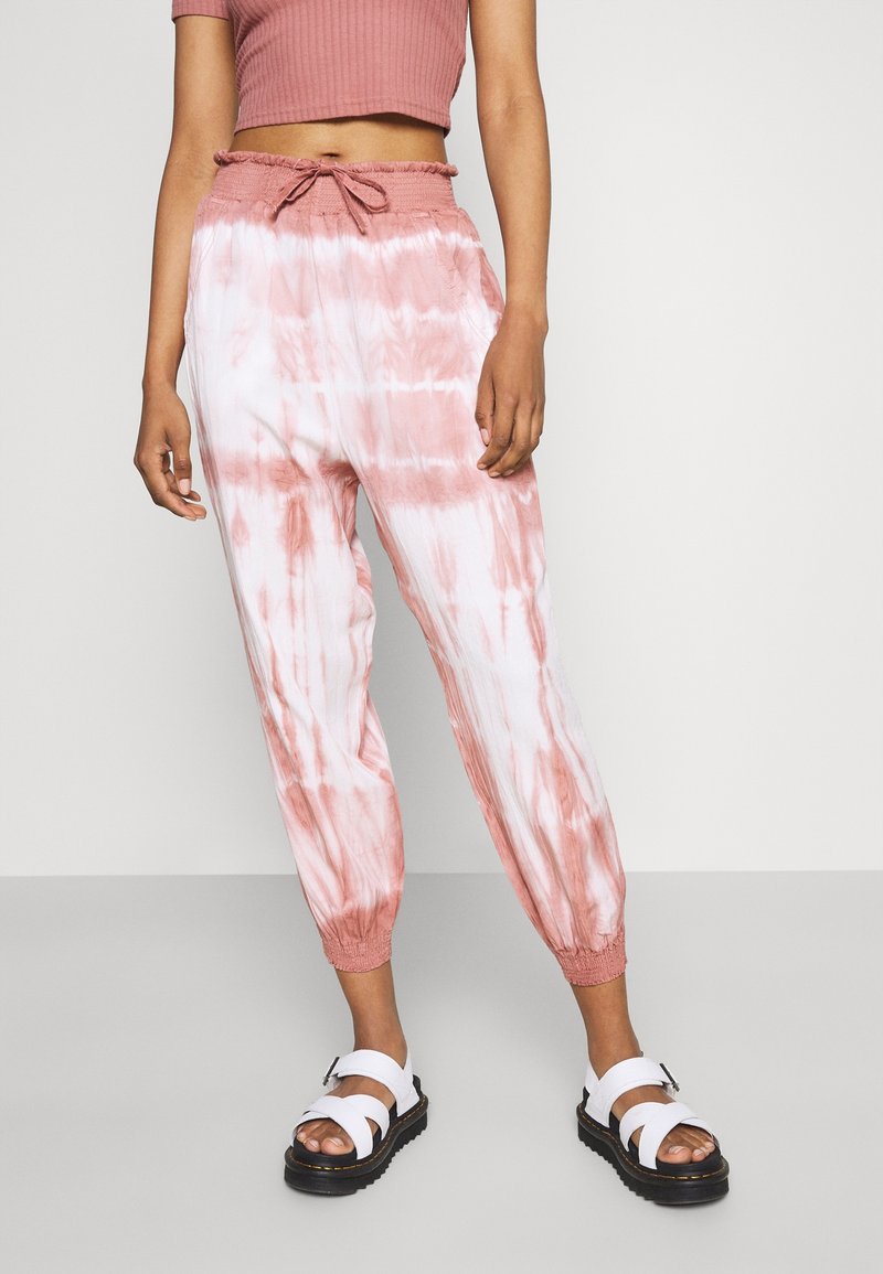 Femme portant un pantalon de jogging tie-dye rose et blanc avec une taille à cordon, un top court rose et des sandales blanches avec semelles noires.