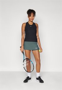 Black sleeveless athletic top, green tennis skirt, black shoes with white accents. Holding a tennis racket and ball. Simple background.