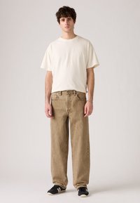 Jonge man staat met een eenvoudig wit oversized t-shirt, ruime beige jeans, zwarte sneakers met witte strepen en een zilveren armband.