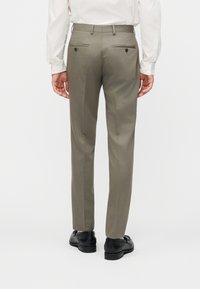 Άνδρας που φοράει χακί παντελόνι με κουμπωμένες πίσω τσέπες, λευκό πουκάμισο και μαύρα παπούτσια, στέκεται με την πλάτη στραμμένη σε λευκό φόντο.