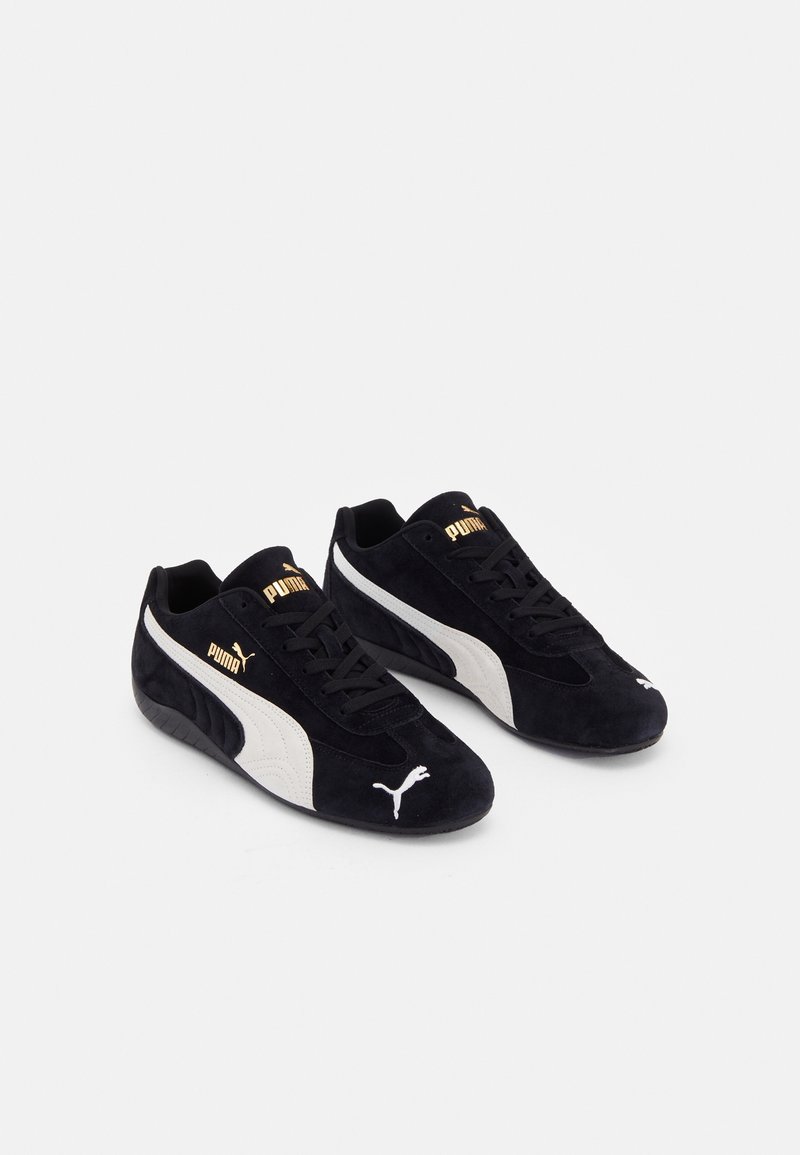 Paire de baskets Puma noires avec des bandes blanches sur les côtés, logo Puma doré sur la languette et le côté, et logo chat Puma blanc sur la zone des orteils.