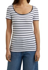 Vrouw draagt een wit T-shirt met korte mouwen en een ronde hals, met marineblauwe horizontale strepen en een blauwe spijkerbroek, staand tegen een effen achtergrond.