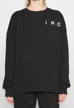 Persoon draagt een zwarte oversized trui met witte letters "IRO" aan de bovenkant links, gecombineerd met een zwarte broek.