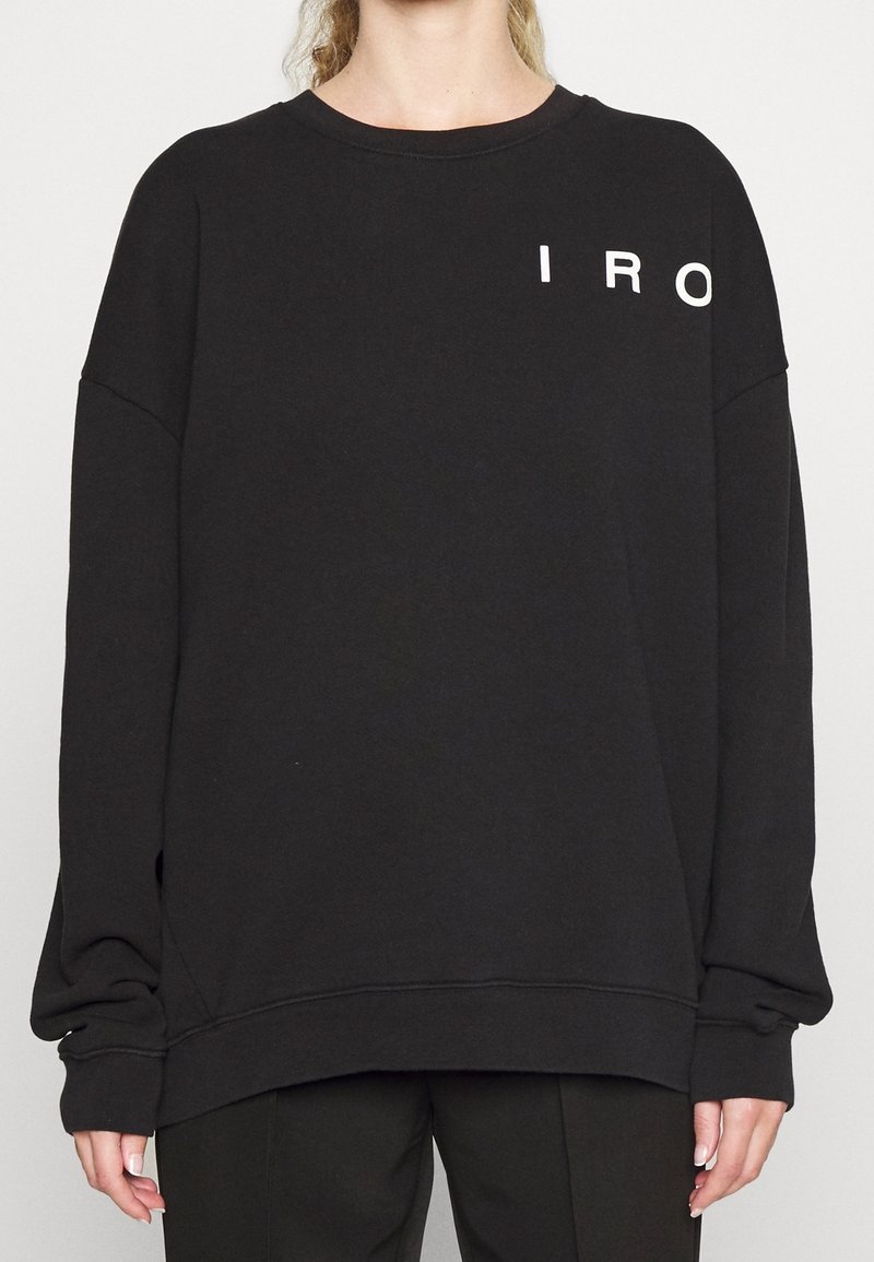 Personne portant un sweat-shirt noir oversize avec des lettres blanches "IRO" en haut à gauche, associé à un pantalon noir.