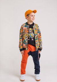 Chłopiec w kolorowej kurtce z graficznymi naszywkami, pomarańczowych i granatowych spodniach, czarnej koszulce z nadrukiem niedźwiedzia, białych sneakersach i pomarańczowej czapce.