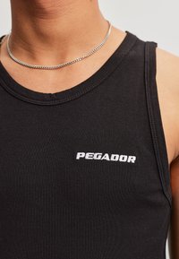 Persoon draagt een zwart geribd hemd met het “PEGADOR”-logo in witte letters en een dunne zilveren ketting om de hals.