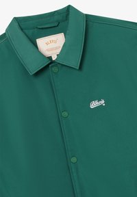 Chemise verte à boutonner avec col, présentant un petit logo blanc "Blkvis" brodé sur la poitrine gauche et une étiquette beige à l'intérieur du col.