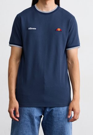 Mann trägt ein marineblaues ellesse T-Shirt mit weiß gestreiftem Kragen und Ärmelsäumen, kombiniert mit blauen Denim-Jeans.