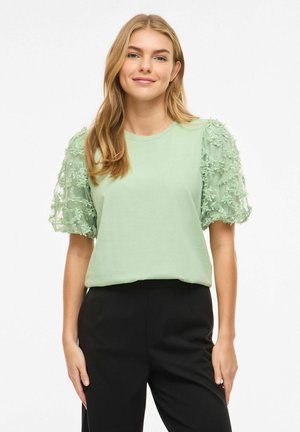 Jeune femme portant un haut vert clair avec des manches bouffantes transparentes à motifs floraux et un pantalon noir, debout devant un fond blanc, souriante.