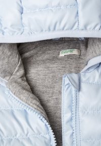 Ljust blått quiltat jacka delvis zipat för att avslöja grå inre foder med en United Colors of Benetton-etikett inuti kragen.