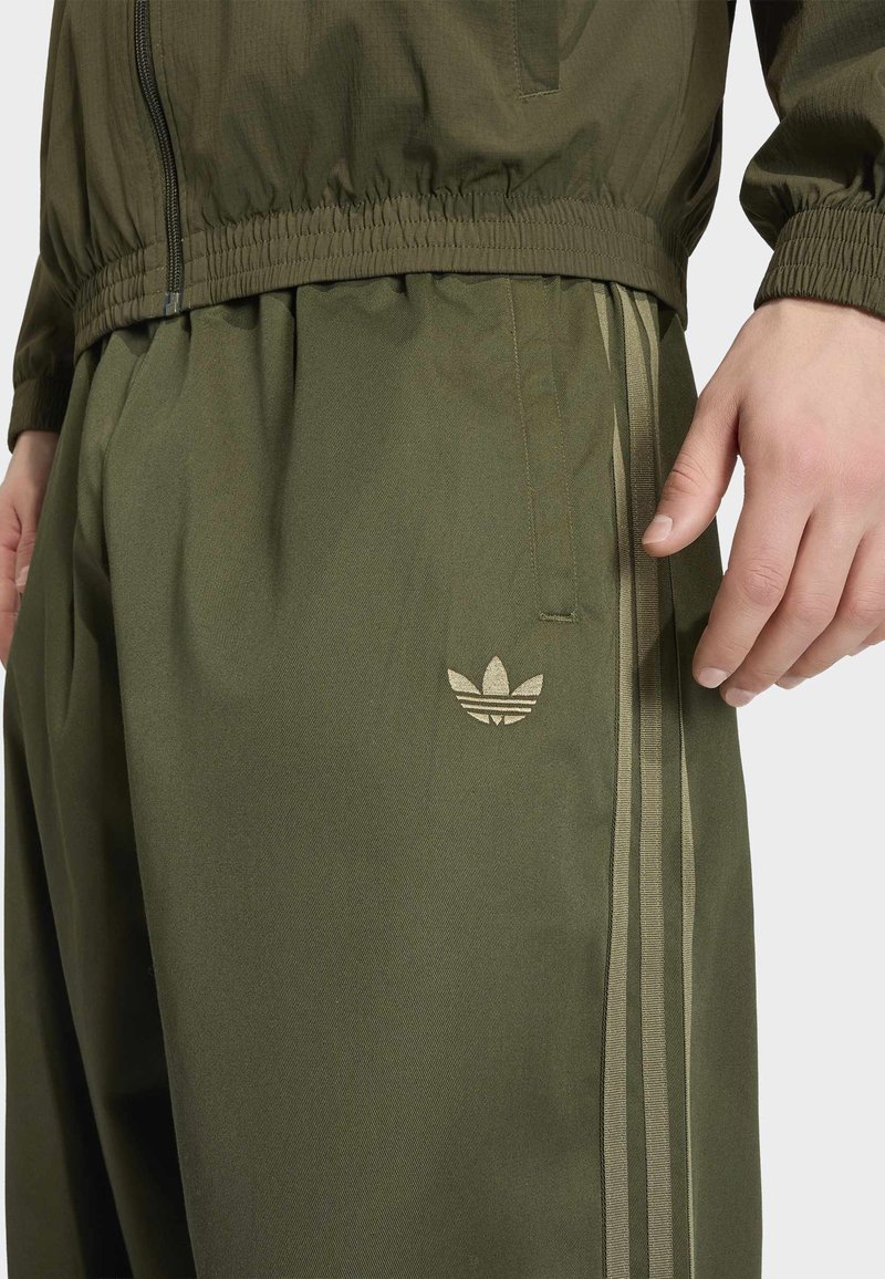 Pantalon de jogging vert olive avec des bandes rayées sur les côtés et le logo Adidas trefoil, porté avec une veste assortie à taille élastique.