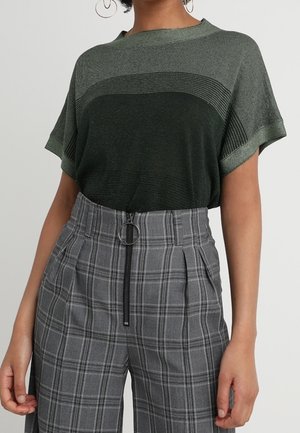 Person iført en grøn strikket kortærmet top og grå ternede bukser med høj talje, frontlynlås og ringtræk.