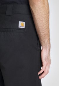 Los pantalones de trabajo negros cuentan con un parche del logo de Carhartt visible en el bolsillo trasero, mostrando un diseño clásico y un material resistente.