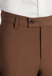 Brązowe eleganckie spodnie o gładkiej fakturze, z zapięciem na guziki i płaskim przodem, zaprojektowane z szlufkami na pasek dla dodatkowej wygody.