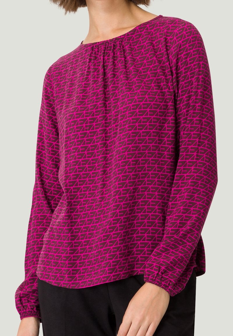 Blouse à manches longues en magenta vif avec un motif géométrique. Dispose d'un col rond et de poignets froncés pour un ajustement décontracté.