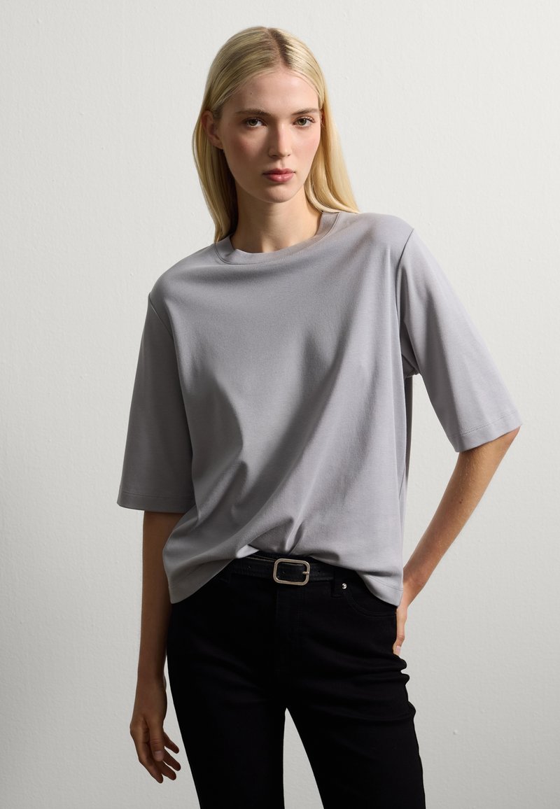 Donna con lunghi capelli biondi che indossa una camicia grigia a maniche corte, larga e infilata in pantaloni neri a vita alta con cintura, in piedi contro un muro bianco.