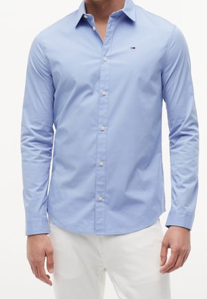 Homme portant une chemise bleu clair boutonnée à manches longues et un pantalon blanc, debout, les bras détendus le long du corps.