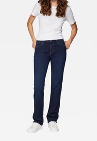 Jeans de mezclilla azul oscuro con corte recto, diseño de cinco bolsillos y costuras visibles, combinados con una camiseta blanca ajustada y zapatillas blancas.