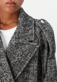 Cappotto in lana houndstooth con un motivo grigio e bianco, ampie rever, due tasche laterali e un bottone metallico decorativo sulla spalla.