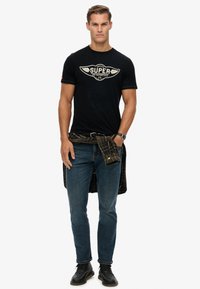 Zwarte katoenen t-shirt met een grafische print met de tekst "SUPER", gecombineerd met blauwe denim jeans en een zwart-groene geruite overhemd om de taille geknoopt.