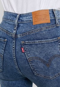 Jeans de mezclilla azul con cintura alta, que presentan una etiqueta de cuero color marrón, cinco bolsillos y un patrón de costura distintivo en el bolsillo trasero.