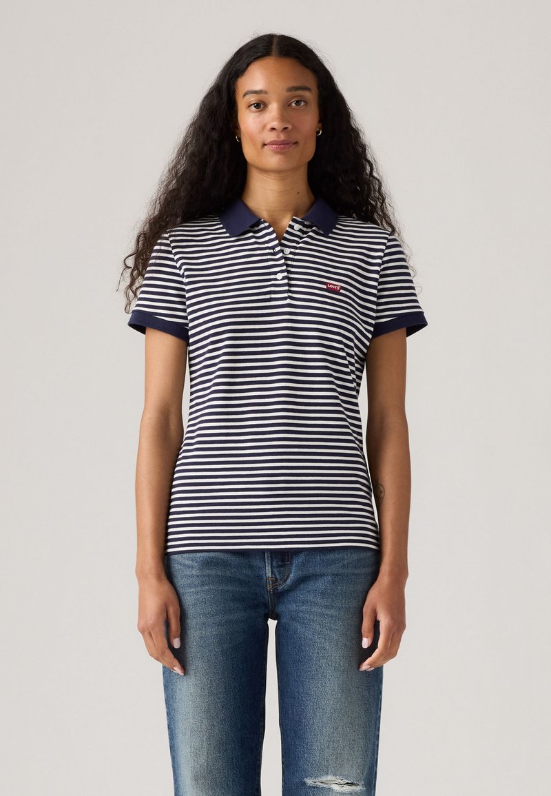 Randig polo skjorta i marinblått och vitt med kort krage och ärmar. Har en liten röd Levi's-logotyp på vänster bröst. Bärs med blå jeans.