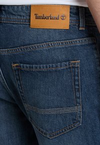 Vaqueros de mezclilla azul oscura con una parche de cuero de la marca Timberland, detalles de doble costura y un bolsillo trasero con acentos naranjas.