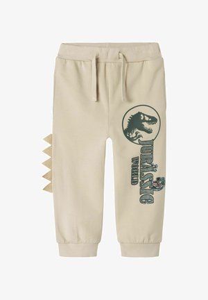 Béžové tepláky se stahovacími šňůrkami v pase, boční grafikou dinosaura a trojúhelníkovými látkovými akcenty na vnější straně nohavic.