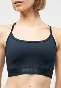 Κοντινή λήψη μιας γυναίκας που φοράει μαύρο αθλητικό σουτιέν με λεπτές τιράντες και ελαστική ταινία κάτω από το στήθος με το λογότυπο MK.